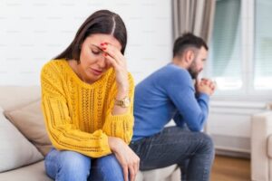 A stressed woman in a yellow sweater sitting on a couch, holding her forehead with one hand, displaying a sense of distress. In the background, a man in a blue sweater sits turned away, indicating emotional distance. This image captures the emotional strain of a couple facing relationship issues, relevant to discussions on divorce and financial disputes.